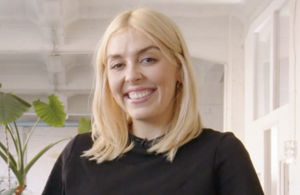 white woman with long blonde hair, which stands in front of a clean grey background looking into the camera