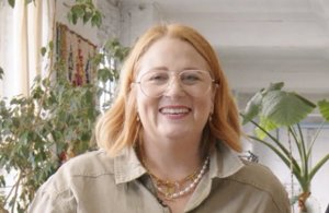 white woman with long blonde hair, which stands in front of a clean grey background looking into the camera