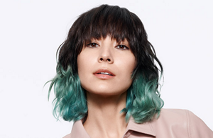 Woman with dark hair, turquoise dyed hair tips and bangs in front of a background.