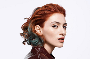 Woman with short, red dyed hair and blue strands of hair in front of a bright background.