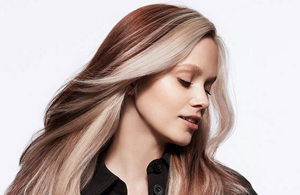 A woman with straight long brown hair with lighter parts close to her face having her eyes closed in front of a bright background.