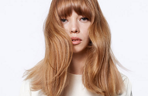 A woman with blonde hair and bangs looking straight into the camera in front of a bright background.