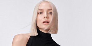A woman with blonde straight hair used elumen dying products on a grey background looks into the camera.