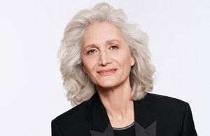 Woman with white hair smiling and glancing at the camera on light gray squared background.