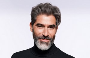 Man with dark grey hair and beard staring at the camera on a light grey  background.