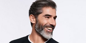 Man with dark grey hair and beard glancing on his left and smiling.