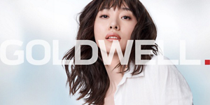Woman with dark wavy hair wearing a white blouse in front of bright background and Goldwell logo in front