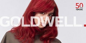 Woman with red hair glancing at the camera on light grey background with the Topchic 50 years logo in the right top corner and the Goldwell logo in the front.