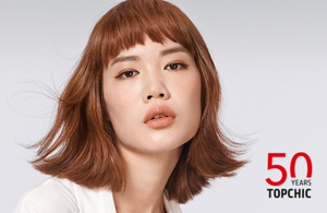 Woman with red hair glancing at the camera on light grey background with the Topchic 50 years logo in the right bottom corner.