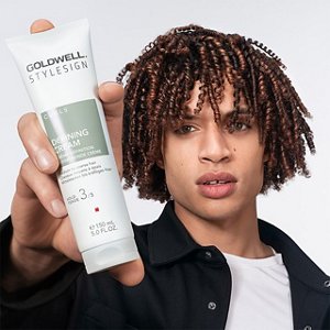a brown haired man with curls and a black jacket is looking straight into the camera while holding a goldwell stylesign curls product in his hands on a white background