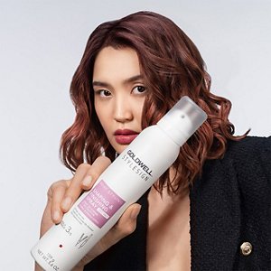 a red haired asian woman with a black blazer is holding a goldwell stylesign heat styling product, while looking in the camera in front of a white background