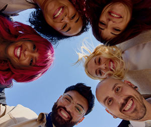  A group of young people in a circle smile down towards the camera with blue sky behind them. From the flowing, bright pink hair to brilliant blond, they showcase the creativity and opportunities available with Goldwell products.