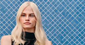 Woman wearing a high neck sleevless satin brown top with her arms crossed standing againsted a blue tiled background with long platinum blonde hair after using Goldwell's Light Dimensions products.