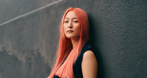 A model stands in front of a dark grey well, showcasing her coral-colored hair for Goldwell's Color of the Year 2026, Bellini.