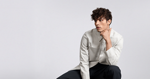 A man sitting down posing in a cream shirt and black jeans with perfeclty curled dark brown hair thanks to using Goldwell's Texture Dimensions to create a curly perm.