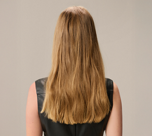 Goldwell Elumen transformation before image showing the back of a model with long, slightly wavy blonde hair against a neutral beige background.