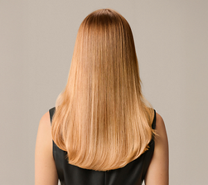 Goldwell Elumen transformation after image showing the back of a model with smooth, glossy, warm blonde hair against a neutral beige background.