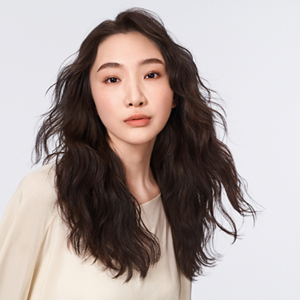 A close up of a woman wearing a beige long- sleeved top with long shiny dark brown wavy hair after using the perm products Texture Dimensions by Goldwell.