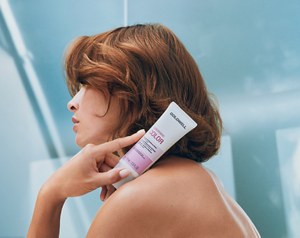 Goldwell Dualsenses Color product held beside a person’s styled auburn hair in a soft blue bathroom setting.