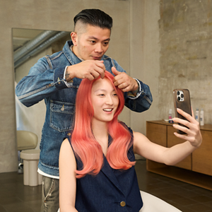 Peter Wu, a Goldwell Global Stylist, points out the vibrance and depth of color in his smiling model's pink hair as she uses her phone as a mirror to look at the finished results. Thanks to Goldwell's product range the model's hair is vibrant and brilliant