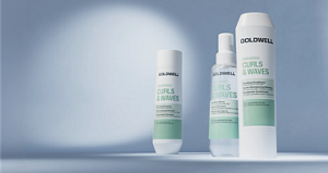 Goldwell Dualsenses Curls & Waves haircare range displayed as white and mint green shampoo, spray and conditioner products on a soft blue studio background.
