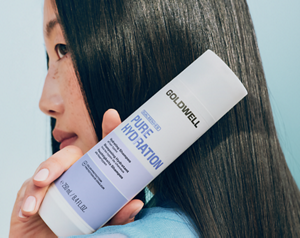 Goldwell Dualsenses Pure Hydration product held beside a person’s long dark hair on a soft blue studio background.
