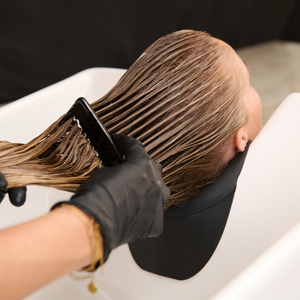 Goldwell Dualsenses image showing gloved hands brushing product through brunette hair at a salon wash basin.