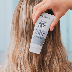 Goldwell Dualsenses Bond Pro image showing a hand holding a white Bond Pro product tube against long blonde hair in a soft salon setting.