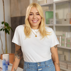 Goldwell Elumen testimonial image showing a smiling blonde woman in a white T-shirt and jeans standing in a bright salon setting. 