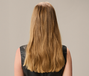 Goldwell Elumen Color transformation before image showing the back of a model with long, slightly wavy blonde hair against a neutral beige background. 
