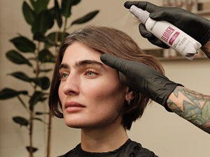 Goldwell Elumen salon application image showing a stylist wearing black gloves applying product from a white bottle to a model with short brunette hair.