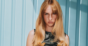 Goldwell hero banner featuring a model with long blonde hair and bangs wearing a black sleeveless top against a pale blue striped background.