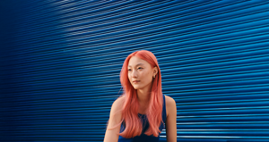 A model in a blue top sits in front of a bright blue background with her vibrant coral-colored hair standing out thanks to Goldwell's Color of the Year 2026 - Bellini