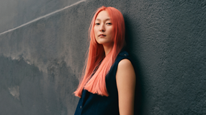 A model stands in front of a dark grey well, showcasing her coral-colored hair for Goldwell's Color of the Year 2026, Bellini.
