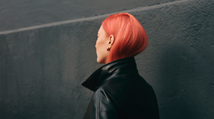 Against her black leather jacket and the dark wall behind her, a model faces away with her coral-colored stright hair tucked into her color. The vibrancy of her hair stands out, celebrating the luminosity of Goldwell's Color of the Year 2026 - Bellini.