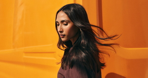 A woman standing against a bright orange background  is wearing a brown dress, with long dark brown shiny hair flowing in the wind, thanks to Goldwell's professional hair salon products.