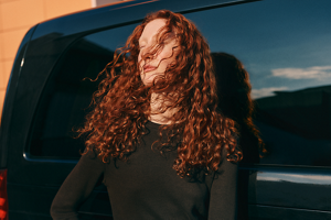 Woman wearing a long sleeved black top standing against a black car with long chestnut red brown curly hair that is healthy and shiny after using Goldwell's Colorance Gloss Toner.