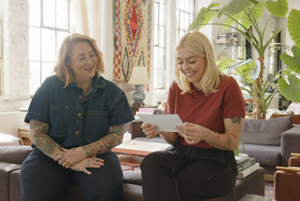 Woman wearing a high neck sleevless satin brown top with her arms crossed standing againsted a blue tiled background with long platinum blonde hair after using Goldwell's Light Dimensions products.