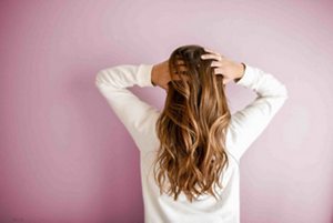 Back of a woman's head reaching for her brunette and blonde highlighted hair against a pink background