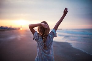 back of a blonde haired girl who is standing on a beach at sunset, wearing a braided hairstyle and lifting her arms up