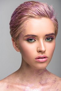 attractive woman with violet glitter on face and hair looking at camera isolated on grey