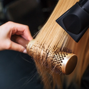 hairdresser blow-drying a woman's blonde fine hair with a hair dryer and a round brush 