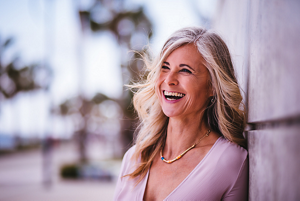 Photo of a woman with grey and blonde wavy hair laughing standing in a street with palm trees