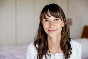 Photo of a woman with long wavy brown hair and fringe sat on a bed smiling wearing a white shirt